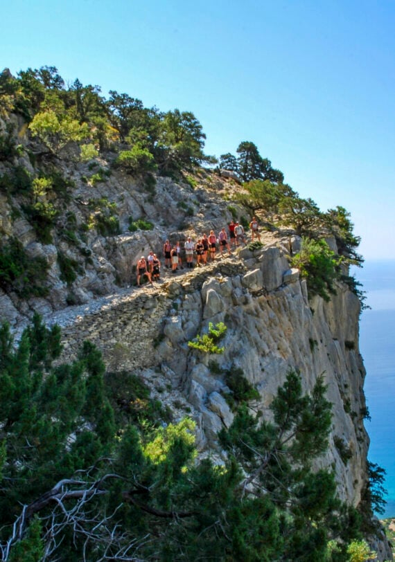 Sentier escarpé dominant la Méditerranée dans les massifs de Sardaigne, en Italie, avec des randonneurs en file indienne à flan de falaise.