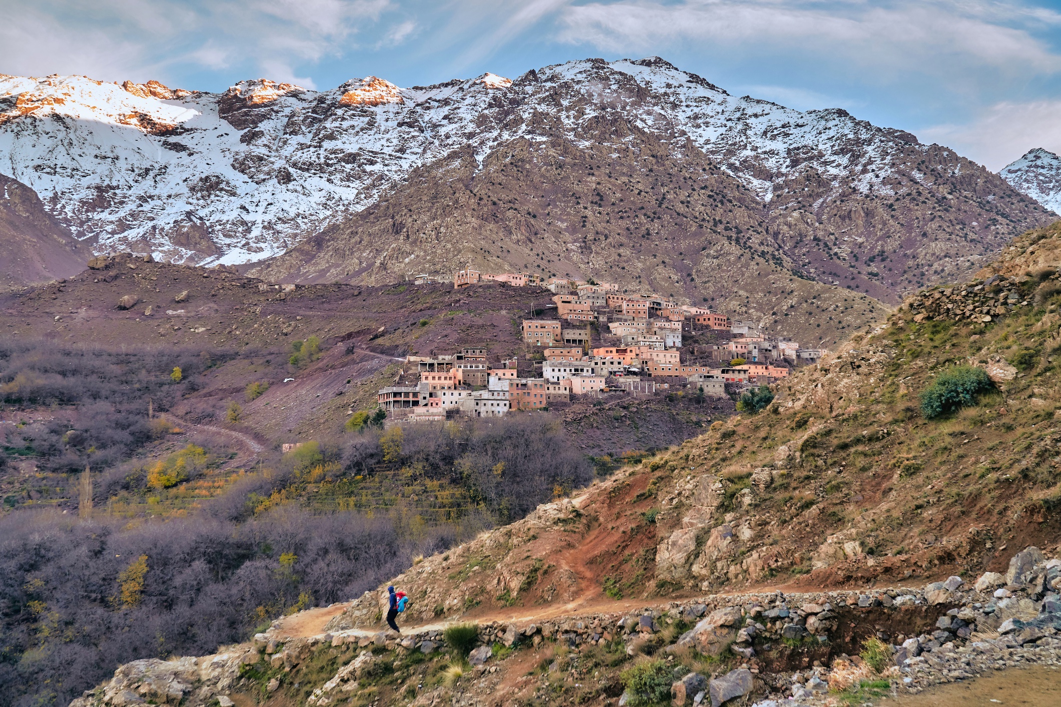  Vue sur un village accroché aux flancs du Haut Atlas marocain, avec des sommets enneigés en arrière-plan et un randonneur au premier plan