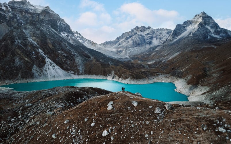 Randonnée vers un lac glaciaire turquoise au Népal. Paysage entouré de sommets enneigés et de reliefs rocailleux en haute altitude, sous un ciel légèrement voilé. Trek avec Karavaniers.