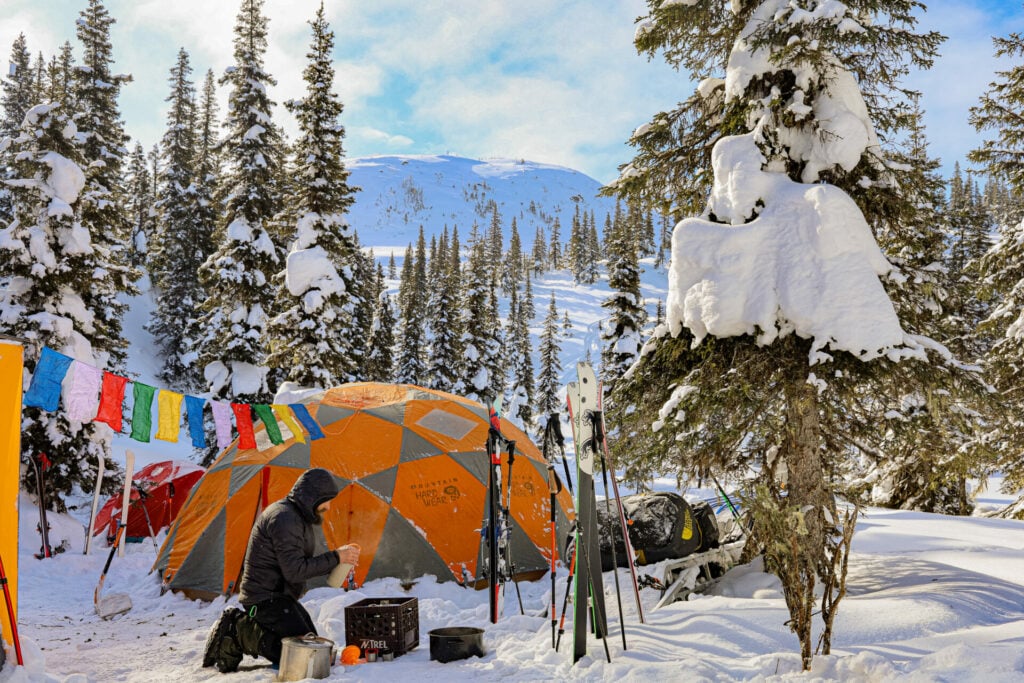Campement hivernal dans les Monts Groulx au Québec, avec tente d’expédition, drapeaux de prière, skis plantés dans la neige et forêt enneigée en arrière-plan.