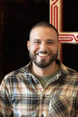 Yassine Temsamani, un homme à la barbe courte et à la coupe rase, portant une chemise à carreaux sur un tee-shirt vert, se tient en souriant devant une porte rouge aux bordures décoratives. - Karavaniers