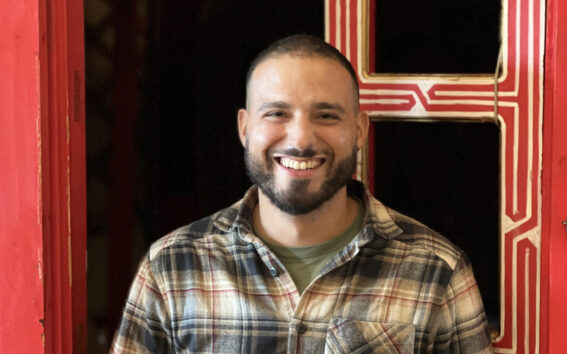 Yassine Temsamani, un homme à la barbe courte et à la coupe rase, portant une chemise à carreaux sur un tee-shirt vert, se tient en souriant devant une porte rouge aux bordures décoratives. - Karavaniers