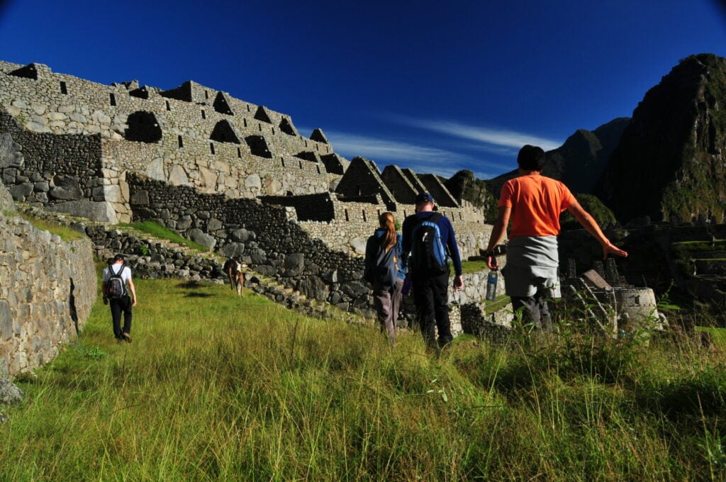 Un groupe de personnes marche dans les ruines herbeuses du Machu Picchu, au Pérou, au cœur de l'Amazonie au Machu Picchu, en direction d'anciens bâtiments en pierre, sous un ciel d'un bleu profond. - Karavaniers