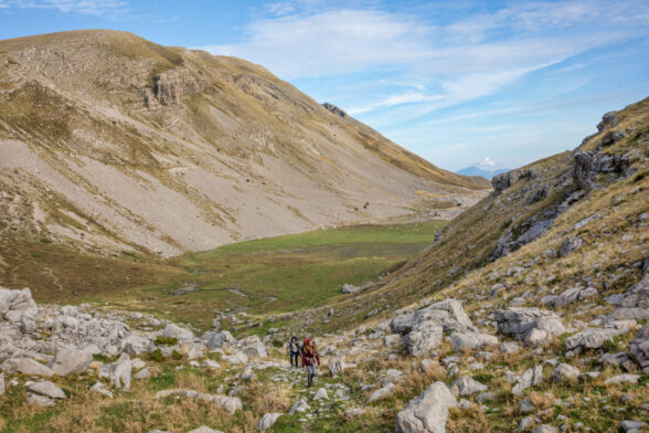 Deux randonneurs et un chien marchent sur un sentier rocailleux dans une large vallée entourée de pentes herbeuses et des spectaculaires montagnes grecques sous un ciel bleu partiellement nuageux. - Karavaniers
