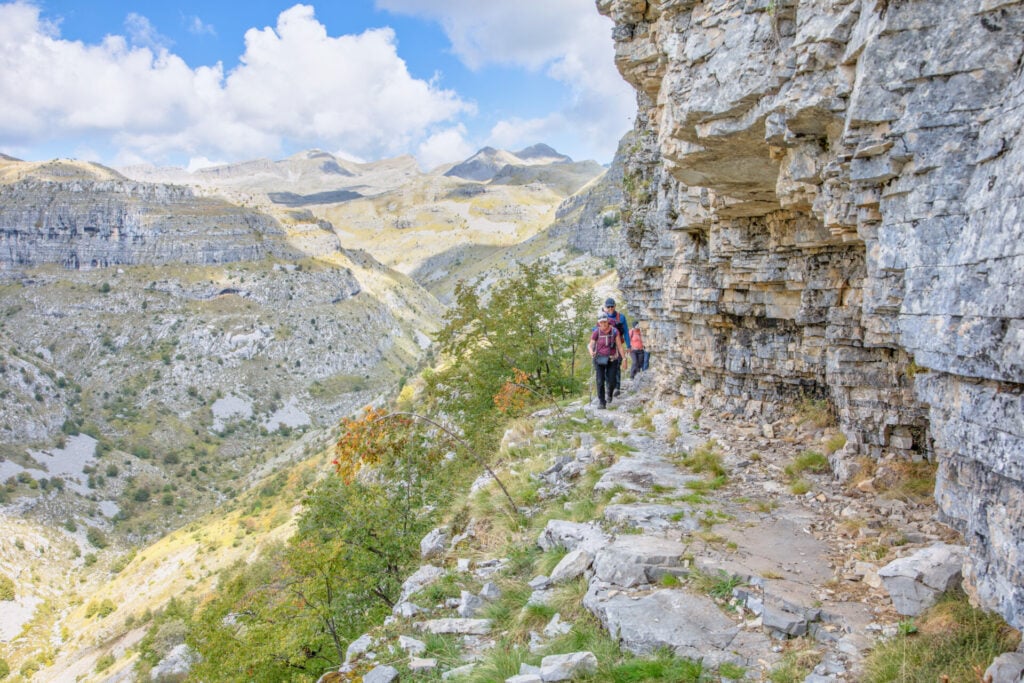 Deux randonneurs marchent sur un étroit sentier rocheux au bord d'une falaise, entourés par les montagnes grecques et un paysage à couper le souffle sous un ciel partiellement nuageux. - Karavaniers