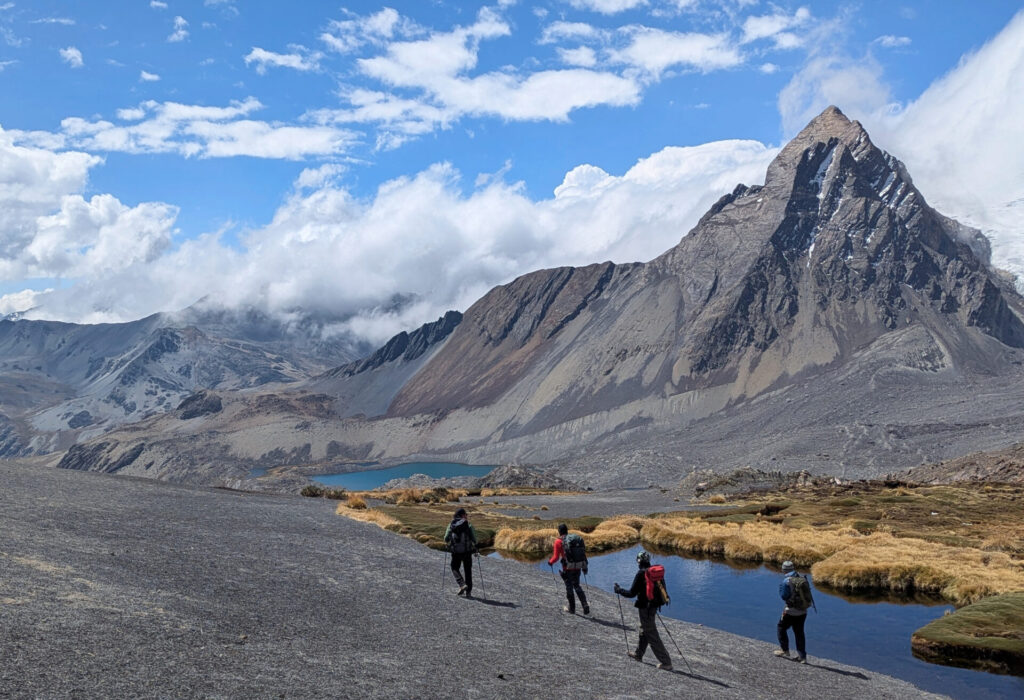 Quatre randonneurs avec des sacs à dos marchent sur un sentier rocailleux et pentu près d'un petit lac, avec en toile de fond les pics acérés de la Cordillère Royale et un ciel partiellement nuageux. - Karavaniers