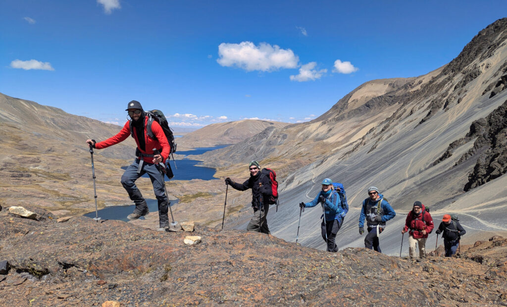 Cinq randonneurs munis de sacs à dos et de bâtons de trekking remontent un sentier de montagne rocailleux le long d'une vallée où se trouve un long lac, entouré des collines escarpées et inclinées de la Cordillère Royale, sous un ciel bleu parsemé de nuages épars. - Karavaniers