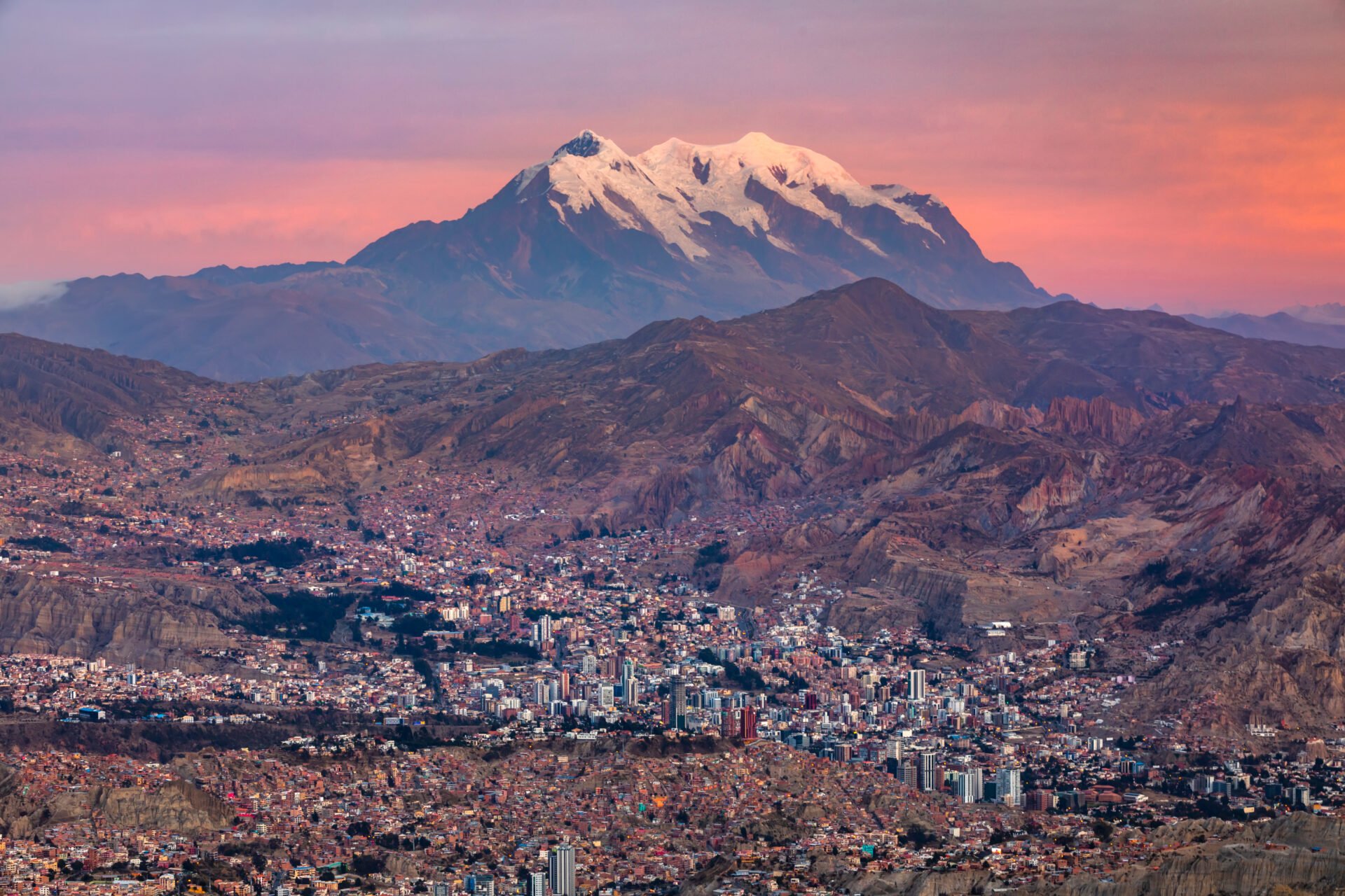 Vue panoramique de La Paz au coucher du soleil, avec ses bâtiments colorés nichés dans une vallée entourée de montagnes, dominée par l’imposant sommet enneigé de l’Illimani.