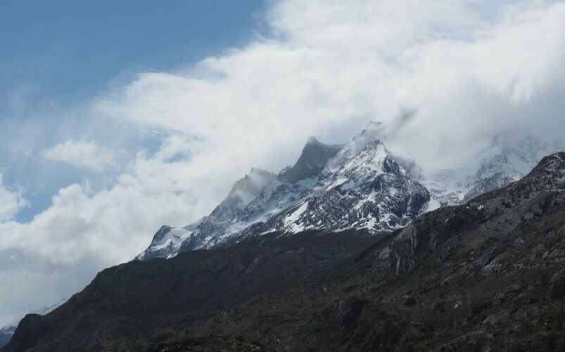 Des pics montagneux enneigés, rappelant les Torres del Paine, s'élèvent au-dessus des nuages avec des pentes rocheuses et herbeuses au premier plan - une scène invitante pour n'importe quelle randonnée sous un ciel bleu clair. - Karavaniers