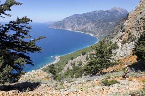 Un randonneur marche sur un sentier rocailleux et pentu à Crète, bordé d'arbres, avec une vue sur la mer turquoise, les collines boisées et les montagnes lointaines sous un ciel bleu clair. - Karavaniers