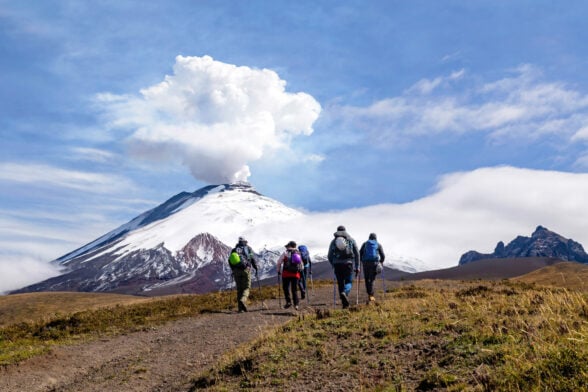 Groupe de randonneurs approchant un volcan actif enneigé, ciel dramatique en toile de fond