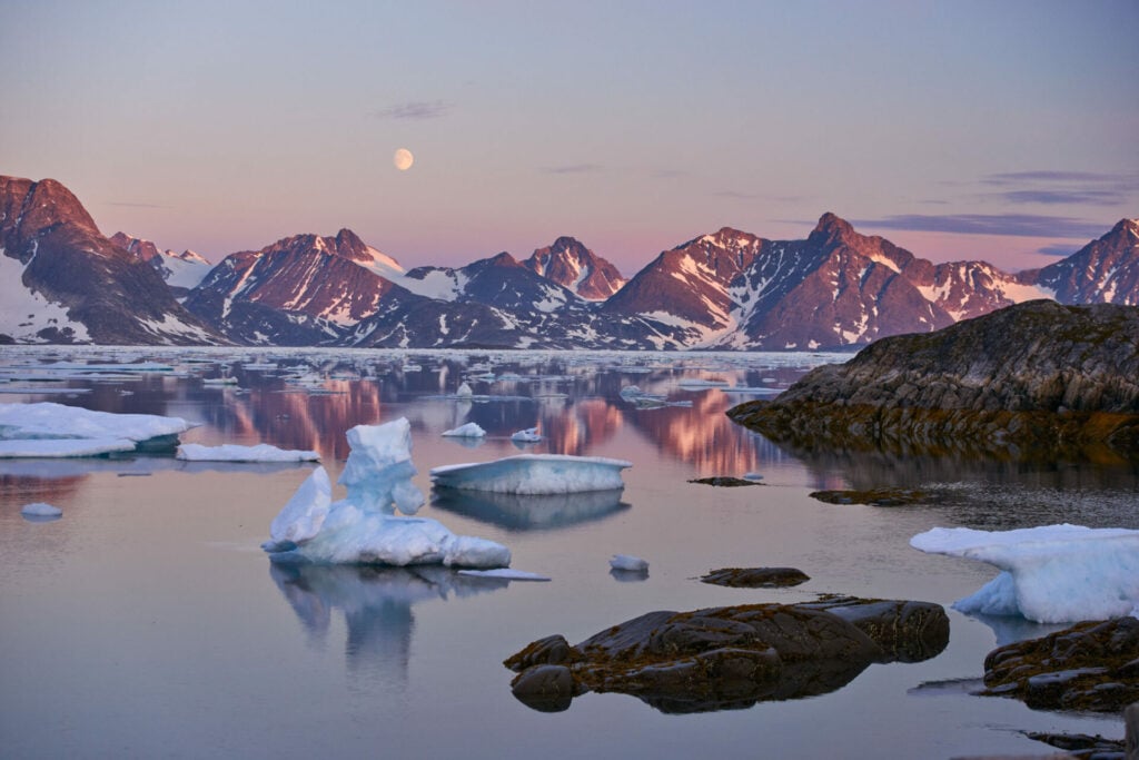 Paysage arctique au Groenland, avec des icebergs flottants sur une mer calme, entourés de montagnes enneigées teintées de lumière rose au coucher du soleil.