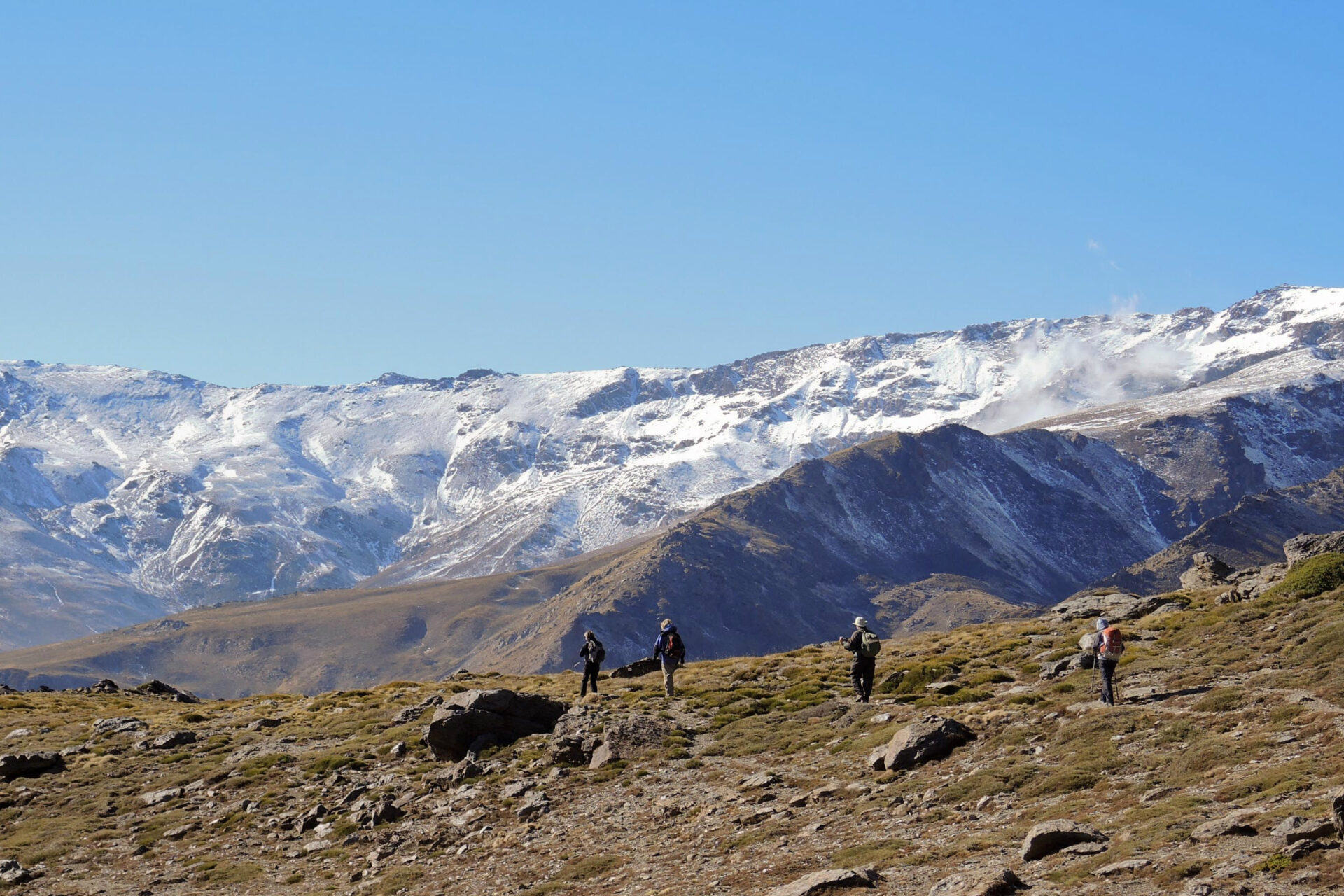 Quatre randonneurs marchent sur un terrain rocailleux à la végétation clairsemée dans les Alpujarras, en direction des sommets enneigés de la Sierra Nevada, sous un ciel bleu limpide. - Karavaniers