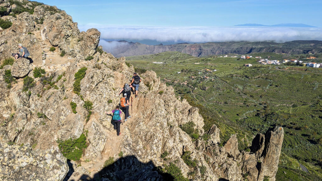 Groupe de randonneurs progressant sur un sentier rocheux en crête à Gran Canaria, avec vue dégagée sur la vallée verdoyante et une mer de nuages à l’horizon.