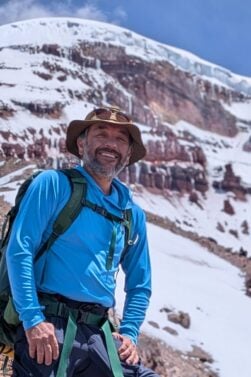Un homme portant une chemise bleue, un chapeau et un sac à dos se tient sur une pente rocheuse avec des montagnes couvertes de neige et de glace en arrière-plan, sous un ciel partiellement nuageux. - Karavaniers