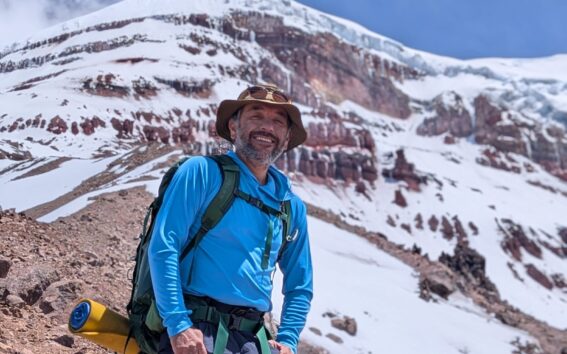 Un homme portant une chemise bleue, un chapeau et un sac à dos se tient sur une pente rocheuse avec des montagnes couvertes de neige et de glace en arrière-plan, sous un ciel partiellement nuageux. - Karavaniers