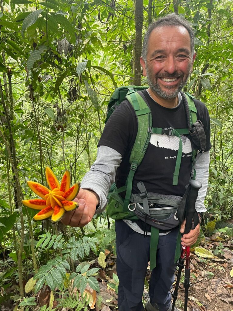 Un homme portant un équipement de randonnée se tient dans une forêt luxuriante, souriant et tenant une grande fleur orange en forme d'étoile avec des taches rouges. Il est entouré d'arbres et d'un feuillage vert dense. - Karavaniers