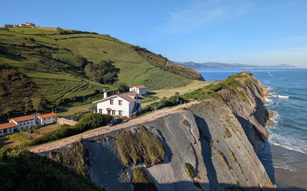 Une maison blanche se dresse sur une falaise surplombant l'Atlantique, avec des collines verdoyantes en arrière-plan et des vagues s'écrasant en contrebas lors d'une journée ensoleillée. Plusieurs voitures sont garées près de la maison dans ce décor pittoresque du Pays basque, avec quelques autres bâtiments à proximité. - Karavaniers
