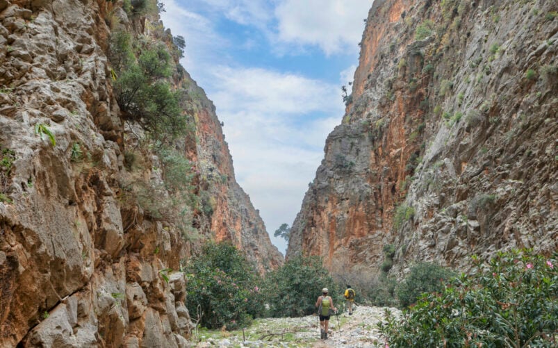 Deux randonneurs profitent de voyages en liberté en marchant sur un sentier rocailleux entre les parois abruptes d'un canyon, sous un ciel partiellement nuageux, entourés de buissons verts et d'une végétation clairsemée. - Karavaniers
