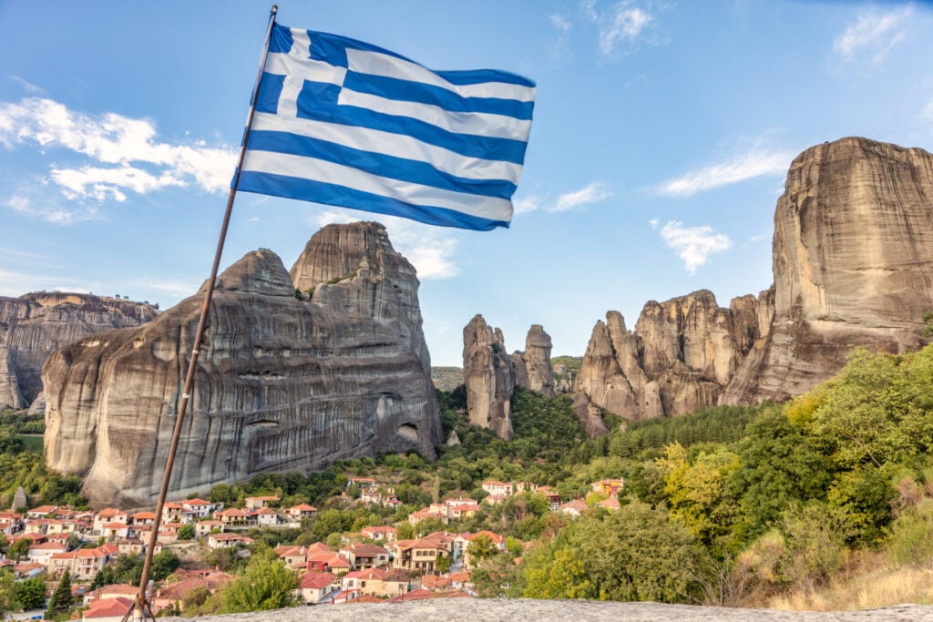 Un drapeau grec flotte au premier plan avec les montagnes grecques des Météores et un village aux maisons à toit rouge entouré de verdure à l'arrière-plan sous un ciel partiellement nuageux. - Karavaniers