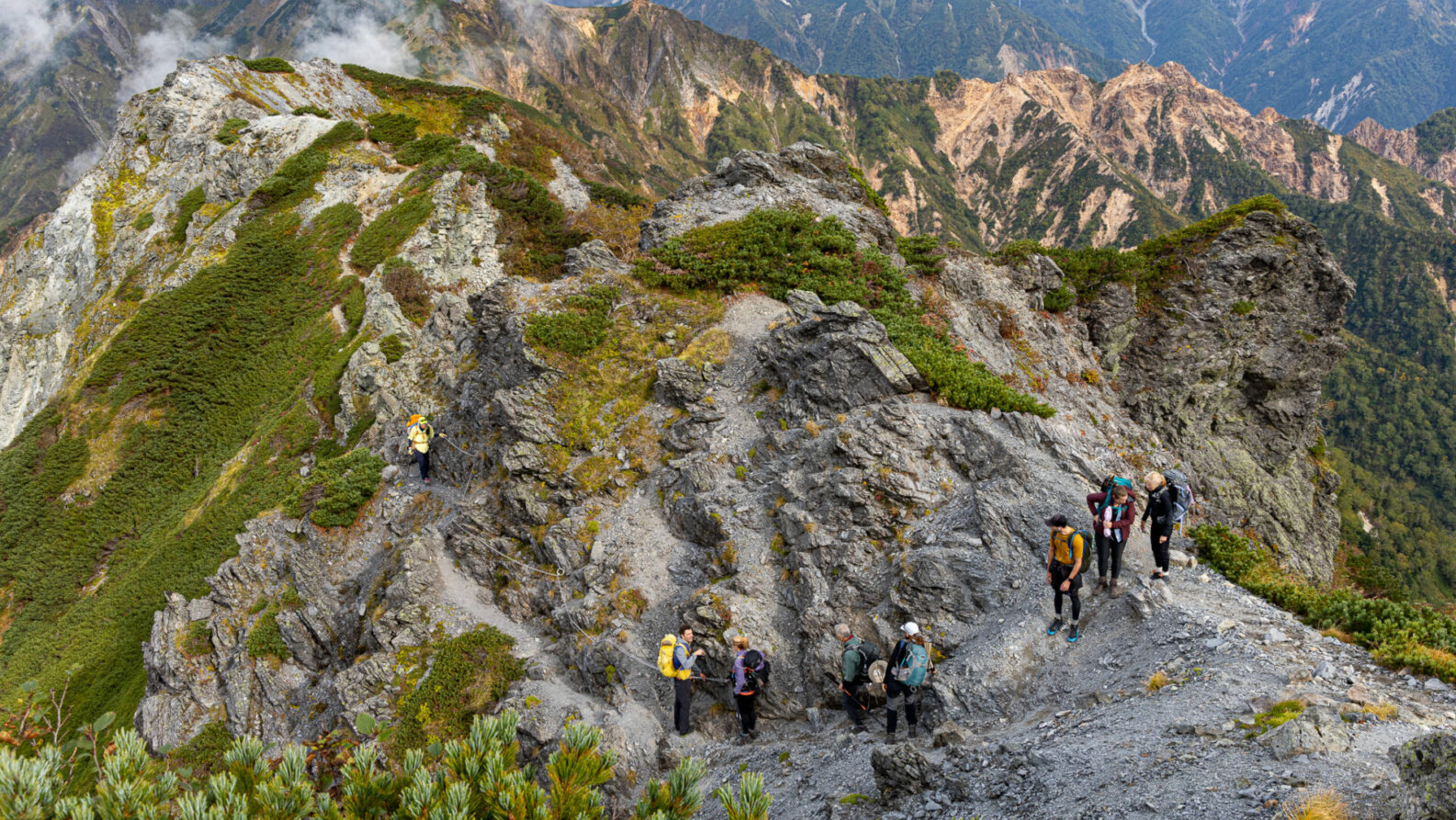 Un groupe de randonneurs portant des sacs à dos marche le long d'un sentier de montagne étroit et rocailleux à Kyushu, au Japon, entouré de pics spectaculaires et d'une végétation verte dense sous un ciel nuageux près de la Mer Intérieure. - Karavaniers