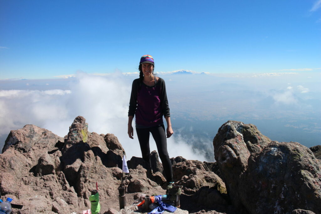 Anie Côté debout sur un sommet rocheux, au-dessus des nuages, sous un ciel bleu éclatant.
