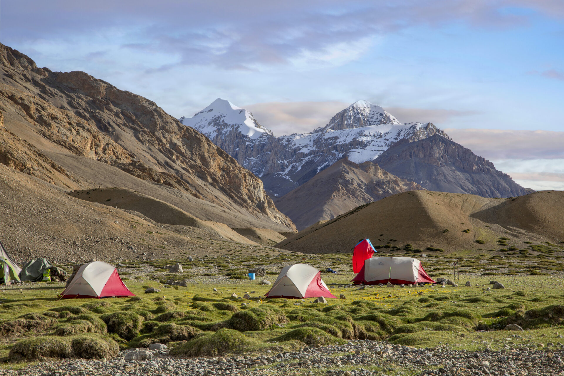 Campement de tentes rouges et blanches installé dans une vallée alpine, entourée de montagnes rocheuses et sommets enneigés sous un ciel légèrement nuageux.