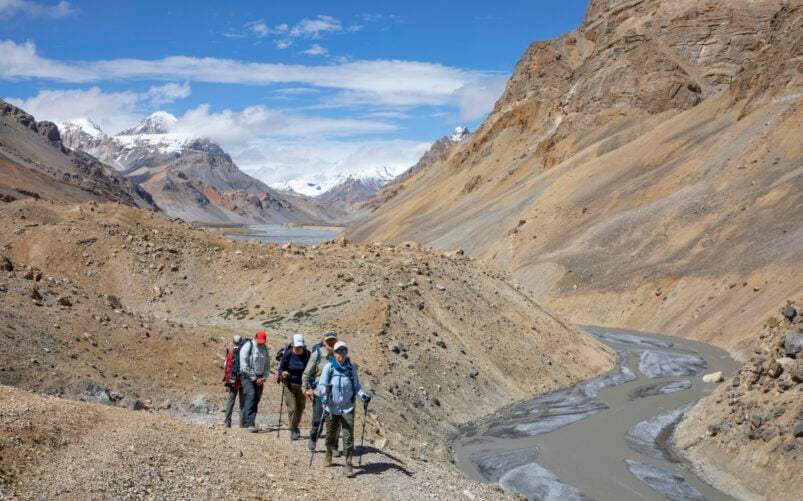 Randonnée dans une vallée aride bordée de montagnes avec un groupe de trekkeurs longeant une rivière sinueuse, sous un ciel bleu dégagé.
