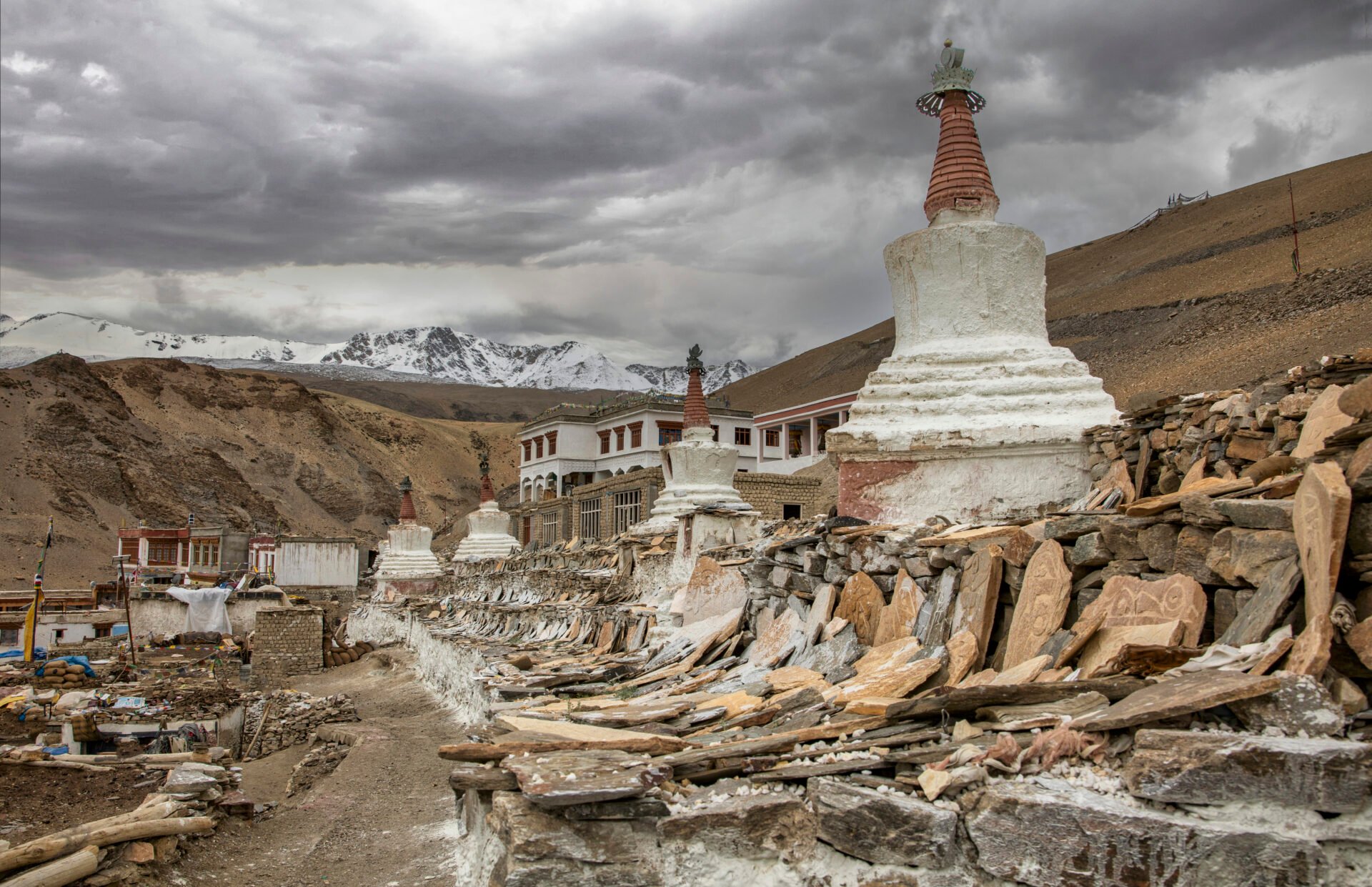 Stupas blancs et murs de pierres gravées dans un village monastique du Ladakh, avec montagnes enneigées en arrière-plan.