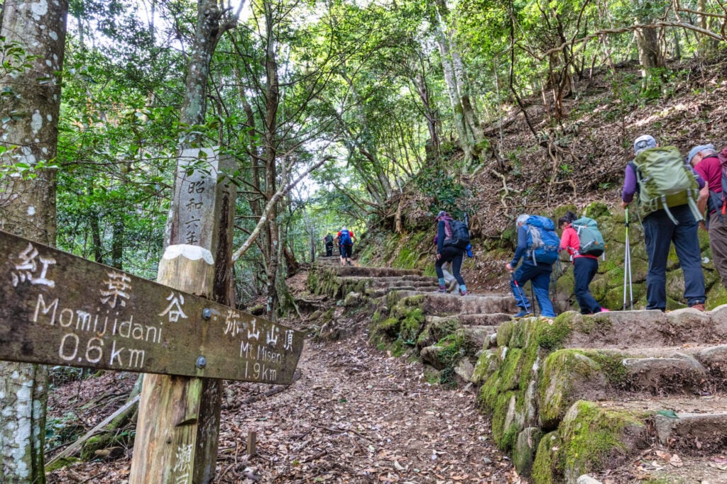 Groupe de randonneurs gravissant un sentier en forêt près d’un panneau indiquant "Mont Iidan".