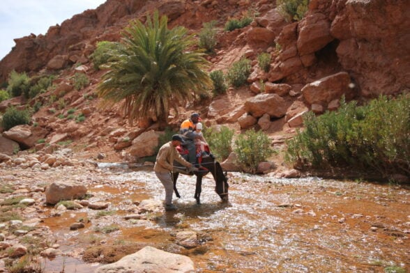 Un homme et un cheval se tiennent dans un ruisseau, tandis qu'un couple de personnes chevauche à proximité - sur fond de beauté sauvage du Massif du Siroua, au Maroc. - Karavaniers