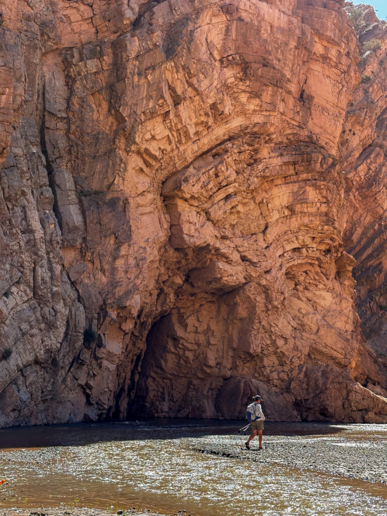 Une personne portant un chapeau et un sac à dos marche le long d'un ruisseau peu profond au pied du grand canyon de roches stratifiées de M'Goun au Maroc, entouré par les falaises abruptes et accidentées de la vallée de la Tessaout. - Karavaniers