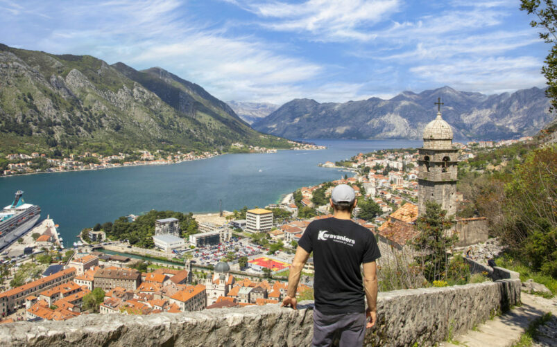 Une personne portant un tee-shirt noir et une casquette se tient sur un chemin de pierre surplombant une ville côtière des Balkans avec des bâtiments aux toits rouges, un bateau de croisière, un clocher d'église et les majestueuses Alpes Dinariques sous un ciel partiellement nuageux. - Karavaniers