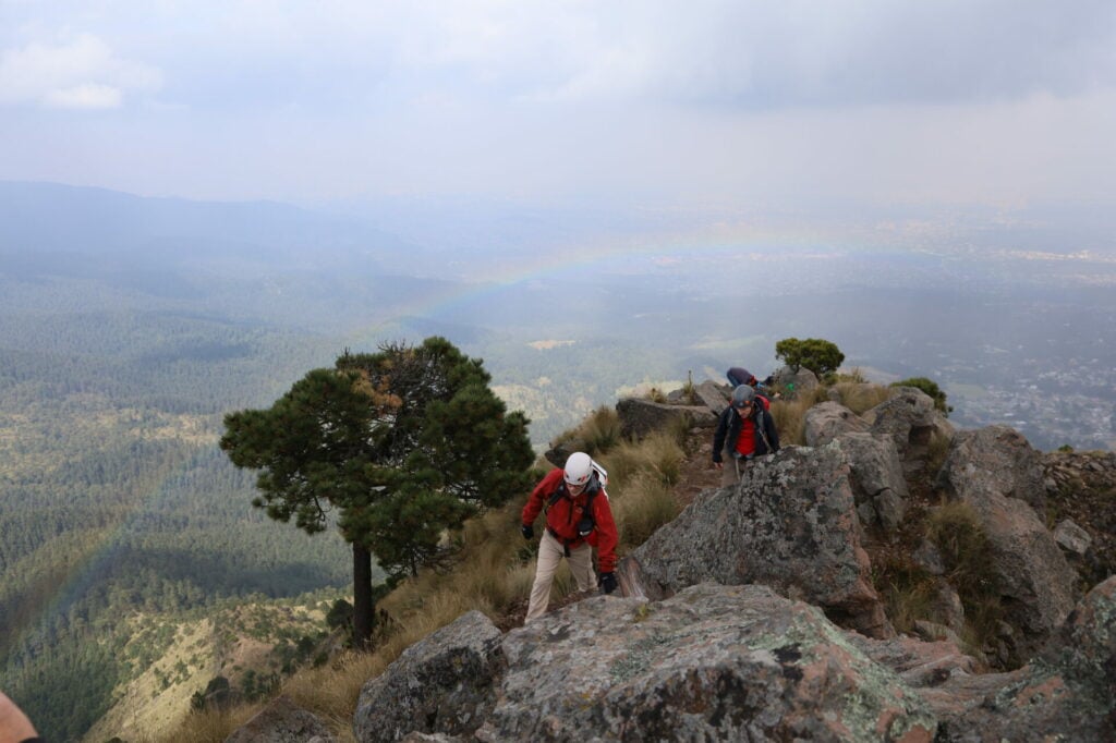 Plusieurs randonneurs portant des casques et des sacs à dos escaladent une crête rocheuse dans la région des Volcans du Mexique, entourée d'arbres. Un léger arc-en-ciel se dessine sur le paysage forestier en contrebas, sous un ciel partiellement nuageux. - Karavaniers