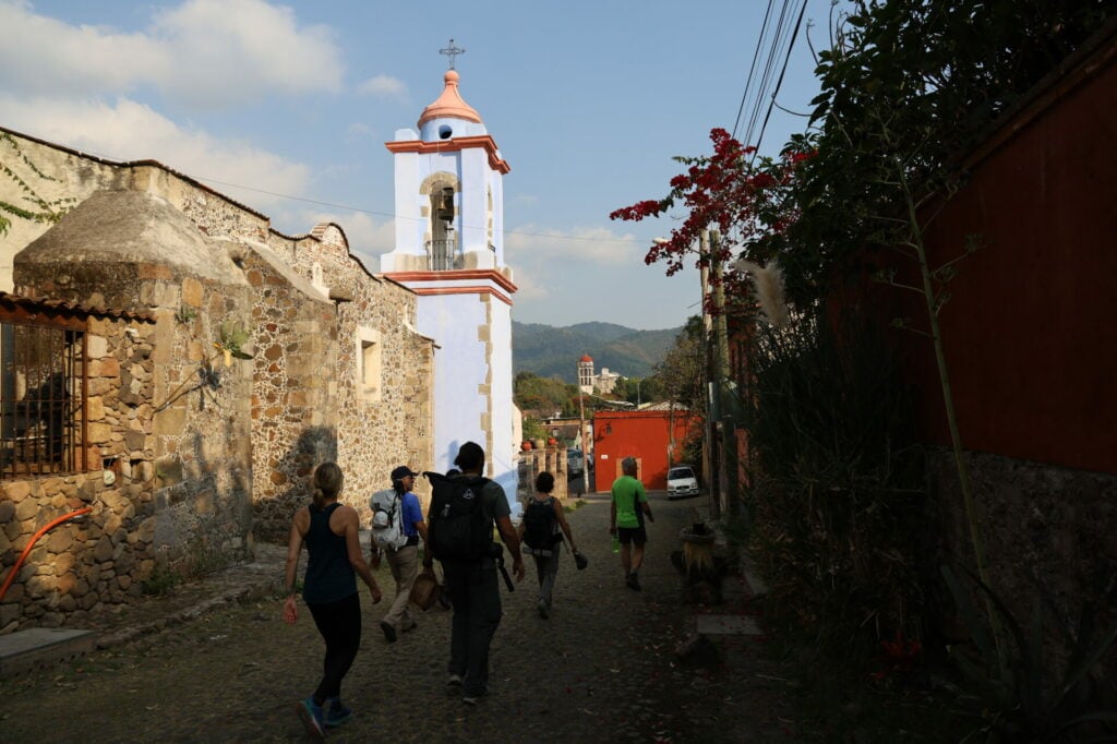 Un groupe de personnes marche dans une rue pavée de l'un des pittoresques Pueblos Mágicos, passant devant des bâtiments en pierre et une église au clocher bleu et blanc ; des montagnes et d'autres bâtiments s'élèvent à l'arrière-plan sous un ciel partiellement nuageux. - Karavaniers