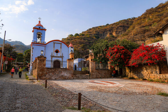 Une petite église à la façade bleue et blanche se dresse devant les Volcans du Mexique. Niché entre les murs de pierre et les fleurs rouges, cet endroit charmant incarne l'esprit des Pueblos Mágicos du Mexique, le long des rues pavées. - Karavaniers