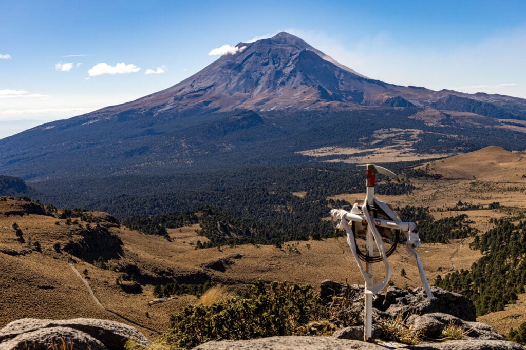 Un instrument géophysique est monté sur des rochers au premier plan, avec en arrière-plan un grand volcan aux sommets enneigés - l'un des emblématiques Volcans du Mexique - et un vaste paysage accidenté sous un ciel bleu limpide. - Karavaniers