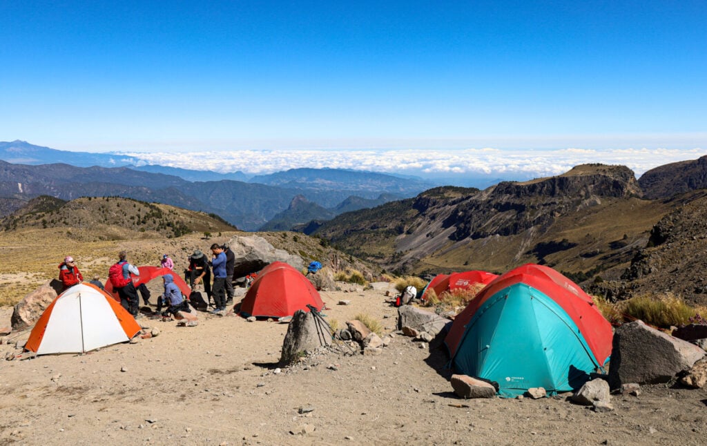 Plusieurs personnes se rassemblent près de tentes dômes colorées installées sur un sol rocailleux dans un camping de montagne, avec des crêtes lointaines et des nuages sous un ciel bleu clair, capturant l'esprit d'aventure que l'on trouve dans la région des volcans du Mexique. - Karavaniers