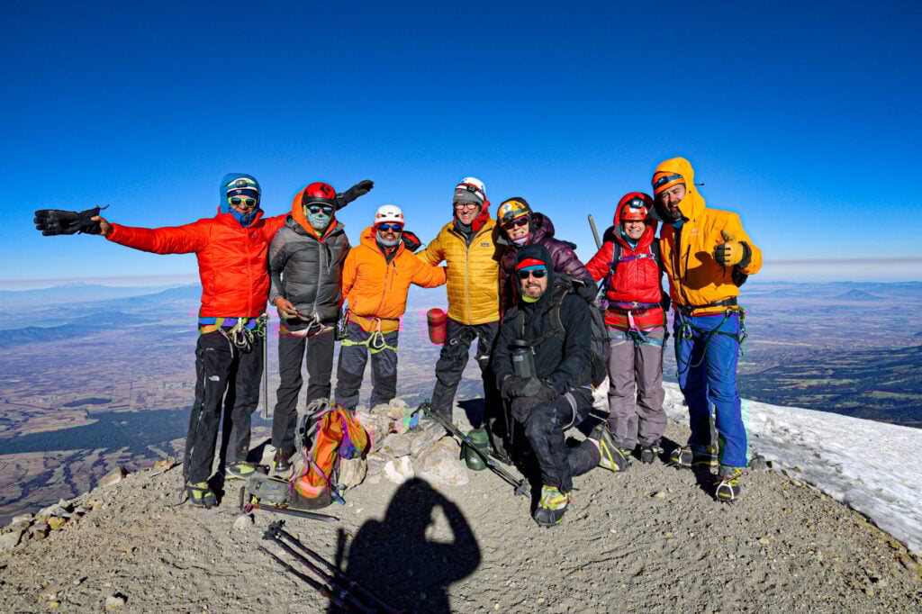 Huit alpinistes portant des casques, des lunettes de soleil et des vestes aux couleurs vives se tiennent ensemble, souriants, sur un sommet des Volcans du Mexique, avec un terrain rocheux et un vaste paysage en arrière-plan, sous un ciel bleu clair. - Karavaniers