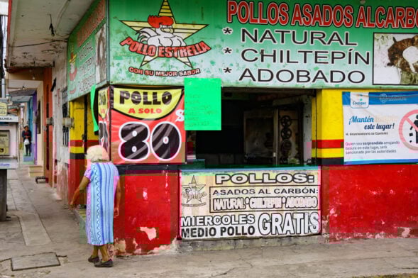 Une femme âgée se tient à l'extérieur d'un magasin de poulets rôtis colorés dans l'un des charmants Pueblos Mágicos du Mexique. Des panneaux peints en espagnol annoncent des poulets grillés, des offres spéciales et des prix. La boutique aux murs rouges, verts et jaunes est animée et dispose d'un comptoir ouvert. - Karavaniers