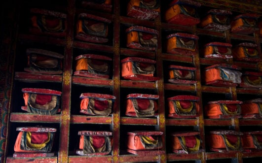 Des étagères remplies d'objets rectangulaires enveloppés et recouverts de tissu rouge et orange, disposés en rangées à l'intérieur d'une structure en bois faiblement éclairée et peinte de façon décorative, le long du Tour du Manaslu, dans le mystique pays de Tsum. - Karavaniers