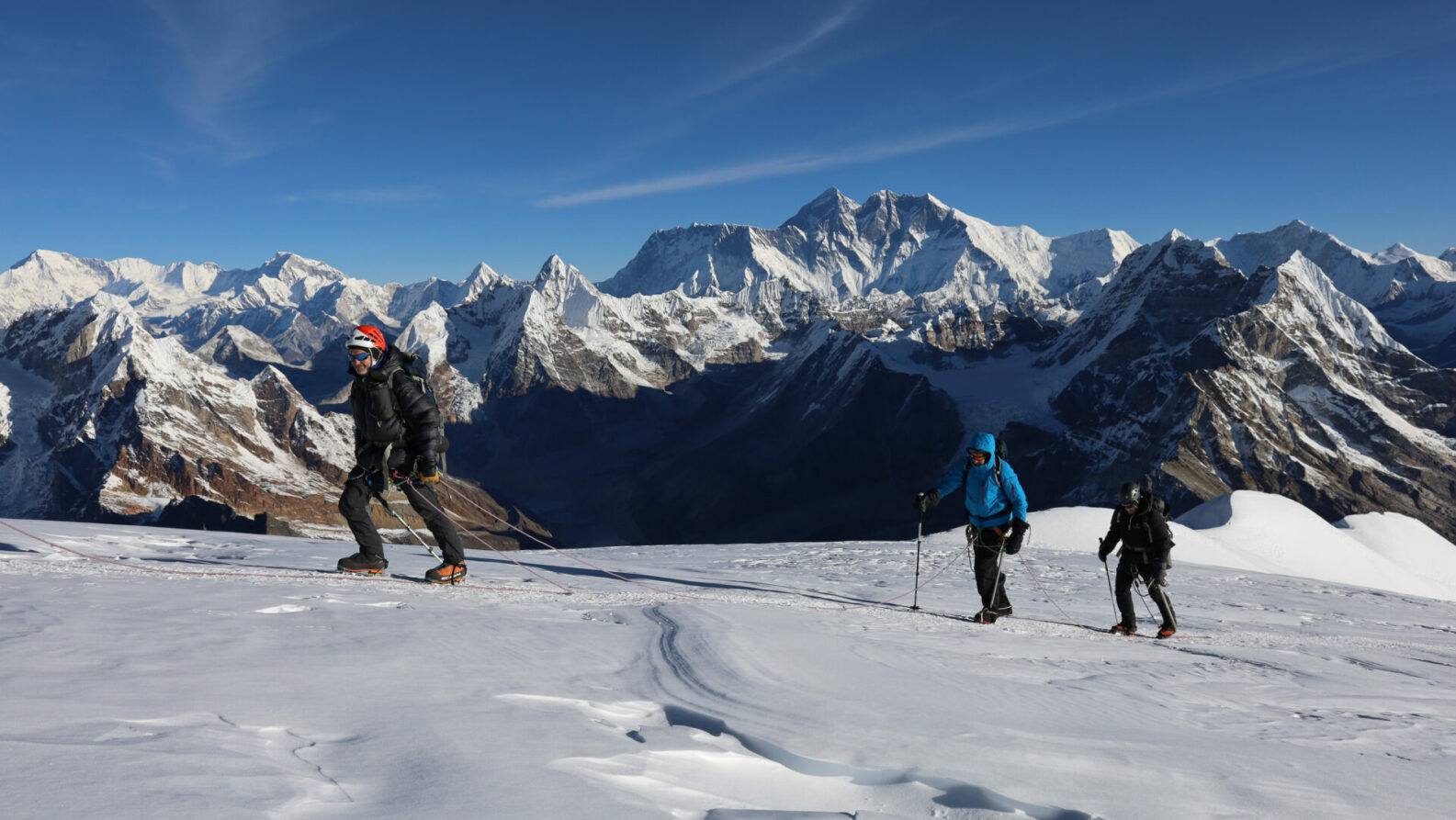 Trois personnes en tenue d'hiver remontent une pente enneigée avec des bâtons de trekking, entourées de montagnes enneigées sous un ciel bleu clair - une aventure qui rappelle les 3 sommets du Kilimandjaro ou du Meru. - Karavaniers