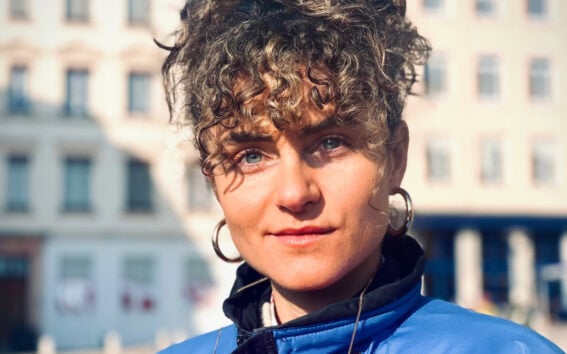 Portrait de Océane Martinez à la lumière naturelle portant un manteau bleu éclatant et de grands anneaux dorés.