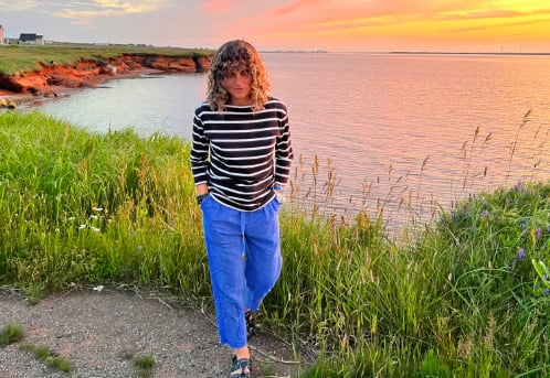 Océane Martinez marchant en bord de mer au coucher du soleil, entre herbes hautes et falaise rouge, dans une lumière dorée.