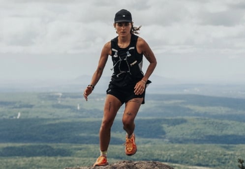 Océane Martinez courant sur un sommet rocheux, vêtue d’une tenue de trail noir et orange, avec une vaste étendue forestière à perte de vue sous un ciel couvert.