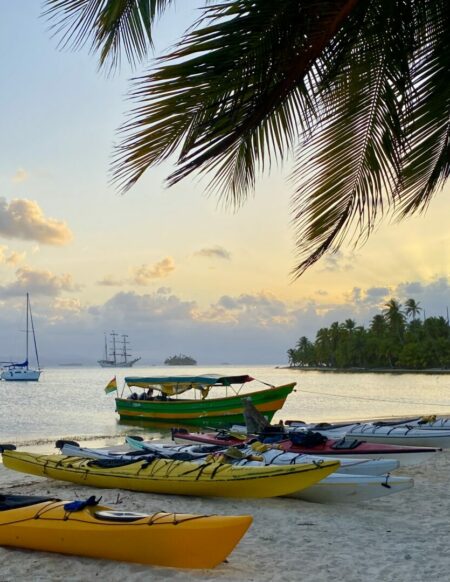Kayaks colorés sur une plage avec palmiers et voiliers au coucher du soleil.