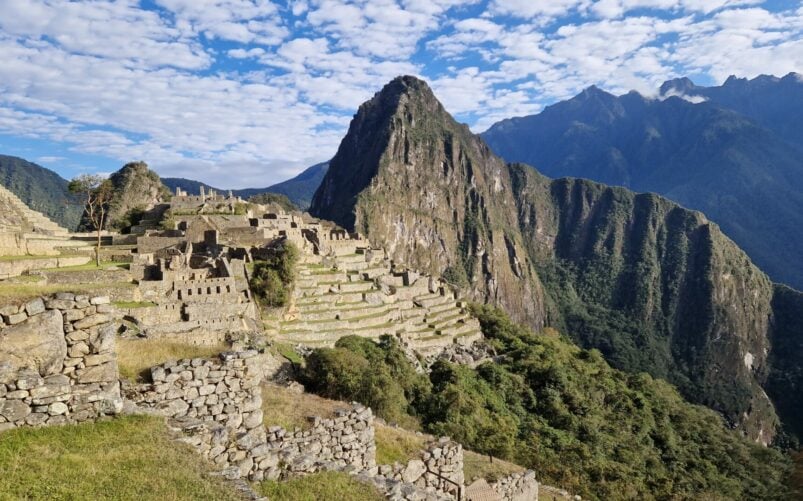 Les anciennes ruines de pierre du Machu Picchu sont situées sur un flanc de montagne en terrasses sous un ciel bleu, avec des sommets verdoyants et la mystique de l'Amazonie au Machu Picchu en arrière-plan. - Karavaniers