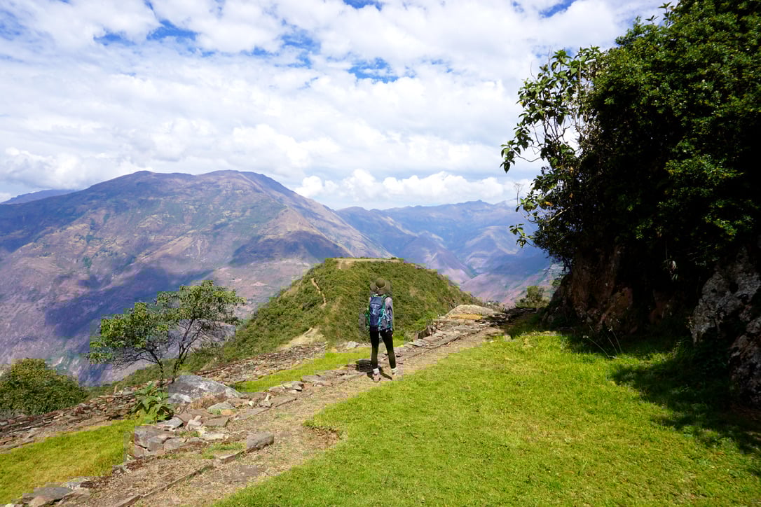 Une personne portant un sac à dos se tient sur un chemin herbeux et rocailleux à Choquequirao, entouré de collines et de montagnes, sous un ciel partiellement nuageux. La végétation verte et le terrain rocheux sont visibles au premier plan. - Karavaniers