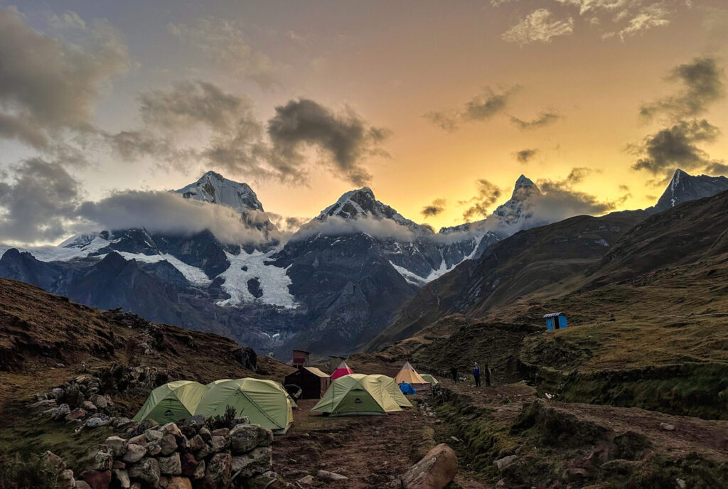 Plusieurs tentes sont installées sur un terrain herbeux le long du Trek du Huayhuash. Les montagnes enneigées brillent au coucher du soleil, les nuages couvrent partiellement les sommets et quelques personnes se tiennent près du campement. - Karavaniers