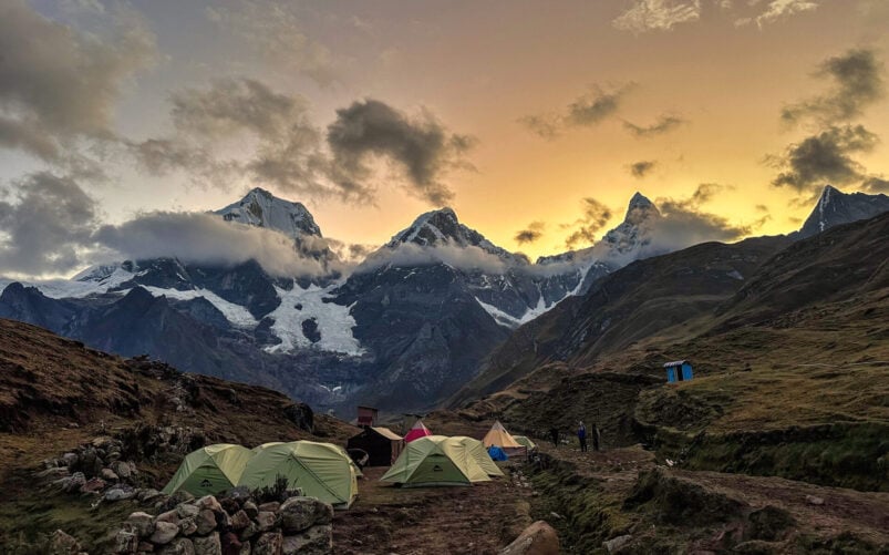 Plusieurs tentes sont installées sur un terrain herbeux le long du Trek du Huayhuash. Les montagnes enneigées brillent au coucher du soleil, les nuages couvrent partiellement les sommets et quelques personnes se tiennent près du campement. - Karavaniers